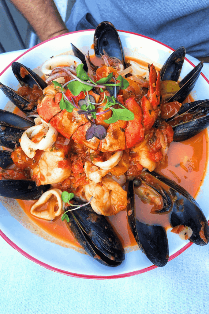 Overhead view of a vibrant seafood dish featuring mussels, lobster, shrimp, squid, and microgreens in a rich tomato broth, served in a white bowl with a red rim.