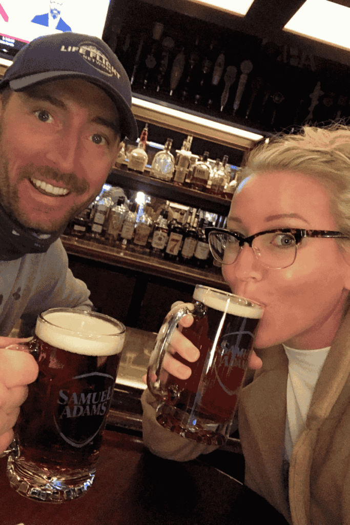 Kate and her husband toasting with Samuel Adams mugs at a cozy bar lined with bottles and beer taps. Classic Boston brews featured on their pub crawl during a weekend getaway in Boston.