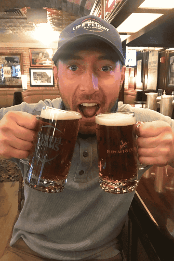 Kate’s husband holding two frosty mugs of beer from Samuel Adams and Elephant & Castle with a playful expression inside a cozy Boston pub. 