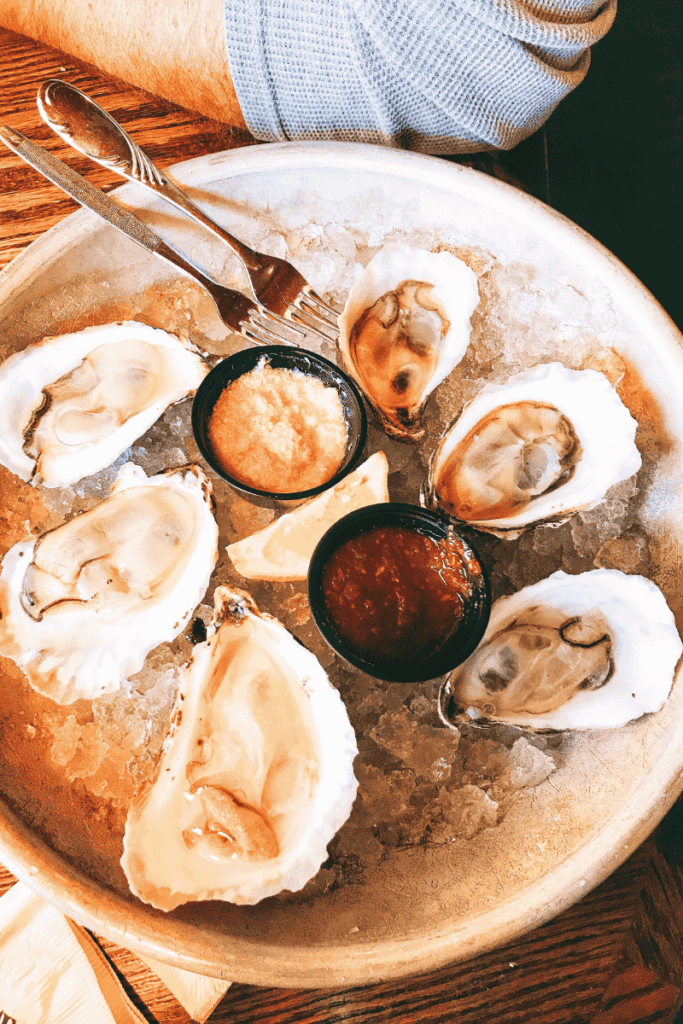 Plate of fresh oysters on ice served with lemon wedges, cocktail sauce, horseradish, and seafood forks on a rustic wooden table.