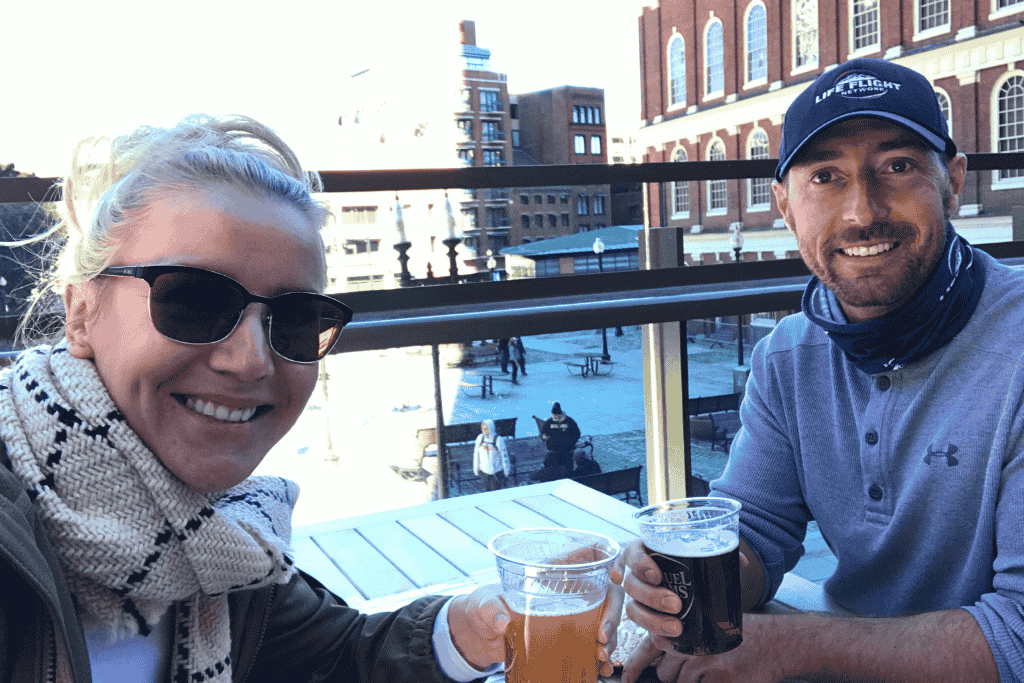 Kate and her husband enjoying beers at an outdoor rooftop table overlooking Faneuil Hall in Boston, both smiling with the historic building in the background. A perfect midday break during their weekend getaway in Boston.
