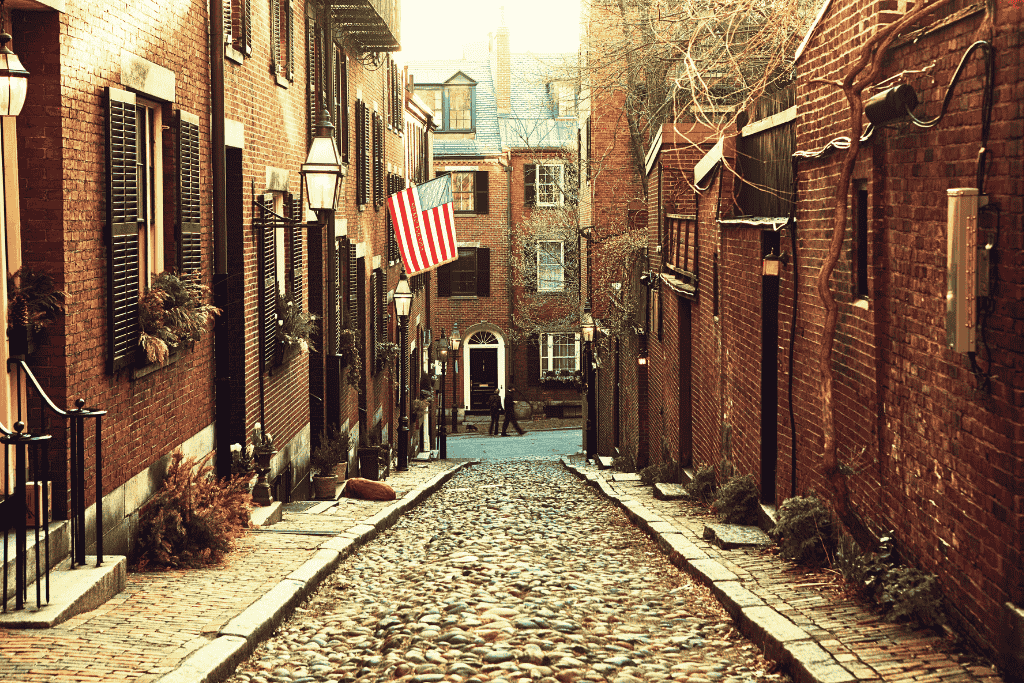 Cobblestone street in Boston’s historic Beacon Hill neighborhood, lined with red brick buildings, black shutters, and an American flag hanging above.