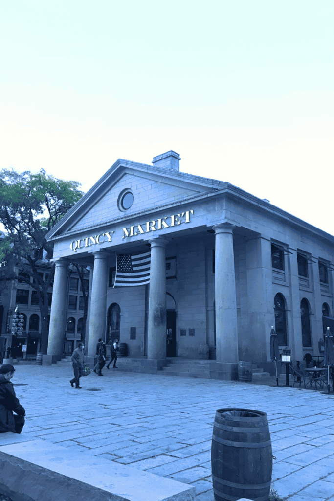 Front view of Quincy Market in Boston with tall stone columns, an American flag, and a few people walking in the plaza.