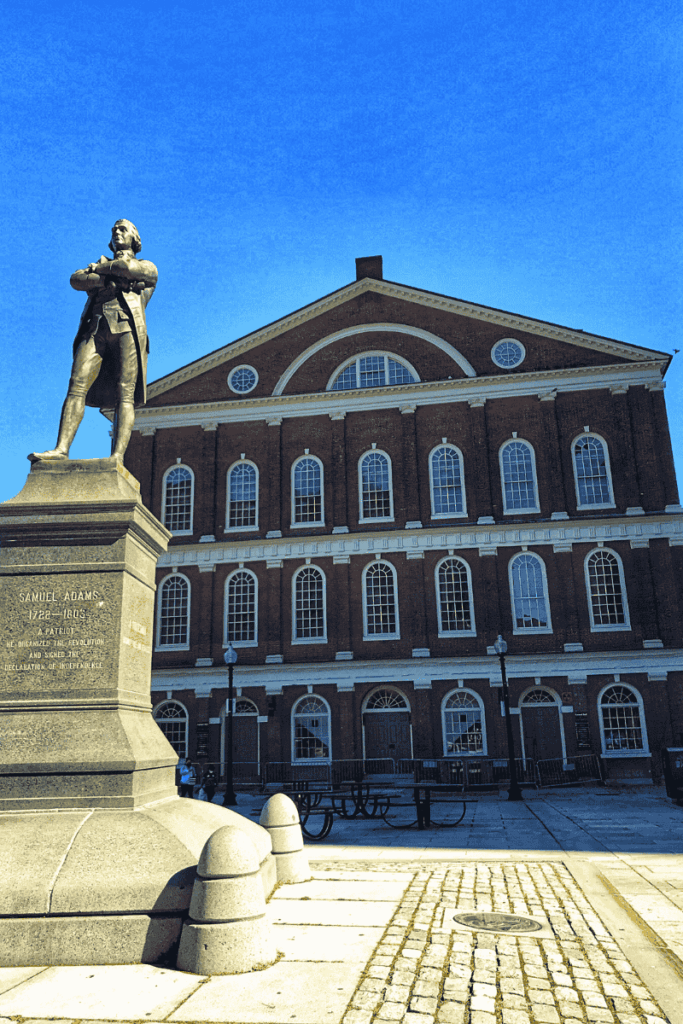 Statue of Samuel Adams standing in front of Faneuil Hall’s historic red brick facade under a clear blue sky.