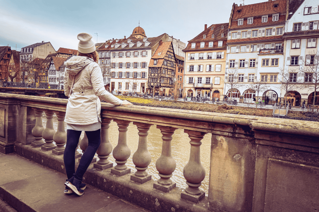 Kate stands at a stone balustrade overlooking a river lined with charming half-timbered and historic buildings in a picturesque European town.