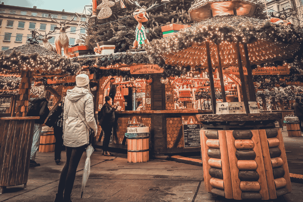 Kate stands with an umbrella in hand, admiring a cozy wooden Christmas market stall decked in twinkling lights, garlands, and playful reindeer figures.