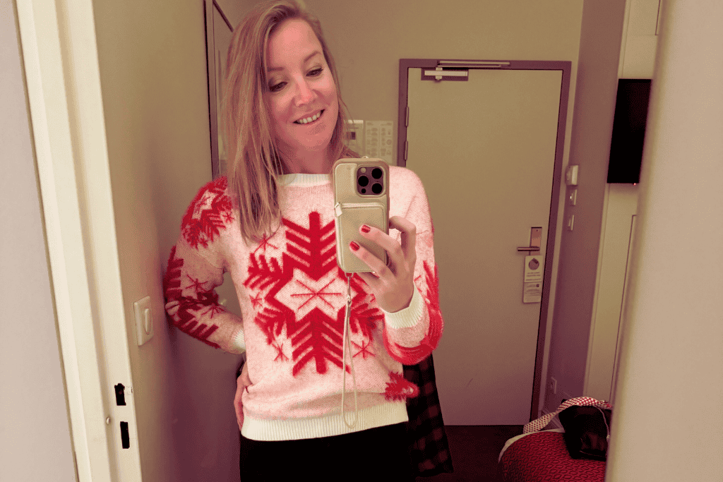 Kate takes a mirror selfie in a hotel room wearing a festive pink and red snowflake sweater, smiling with her phone held up.