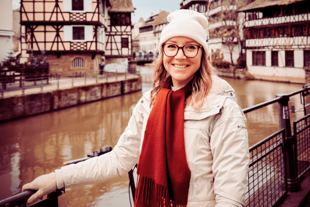 Kate stands along a riverside railing in front of traditional half-timbered buildings, smiling warmly during a winter visit to a European Christmas market.
