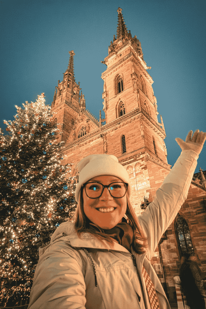 Kate beams in front of a glowing Christmas tree and the illuminated twin towers of Basel Minster, pointing upward in excitement.