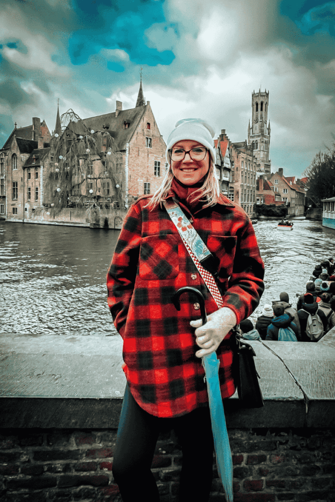 Kate stands beside a canal in Bruges with a smile, holding a blue umbrella and framed by medieval buildings and cloudy skies.