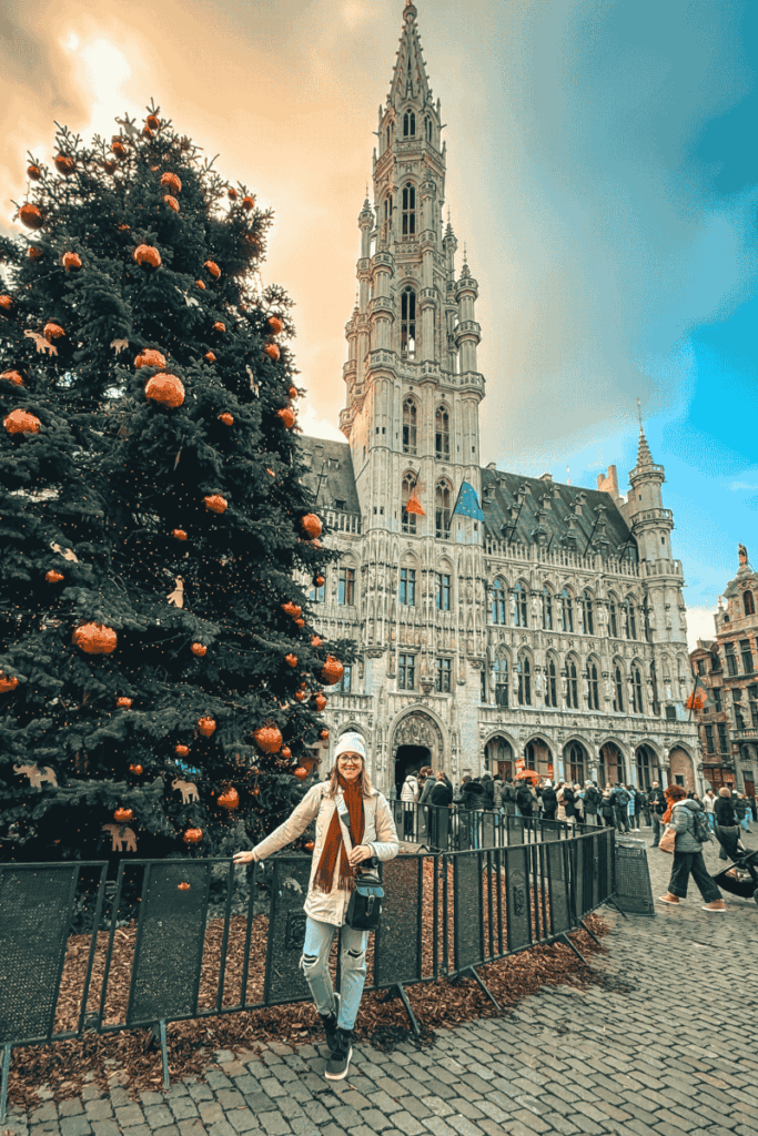 Kate poses next to a large decorated Christmas tree in front of Brussels’ ornate Grand Place town hall with golden light in the background.