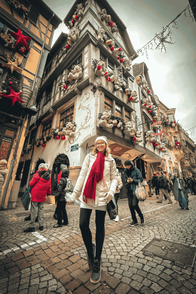 Kate stands smiling on a cobblestone street near a corner building lavishly decorated with teddy bears, ornaments, and festive storefronts.