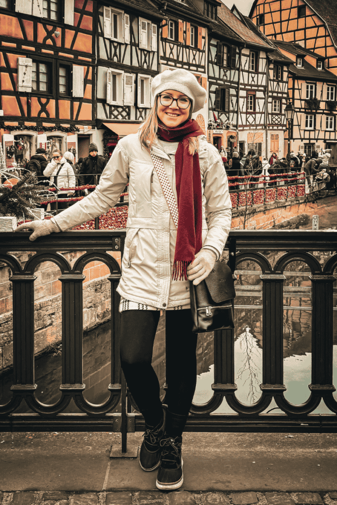 Kate smiles on a bridge in front of colorful half-timbered houses and a canal, bundled up for a winter day at a European Christmas market.