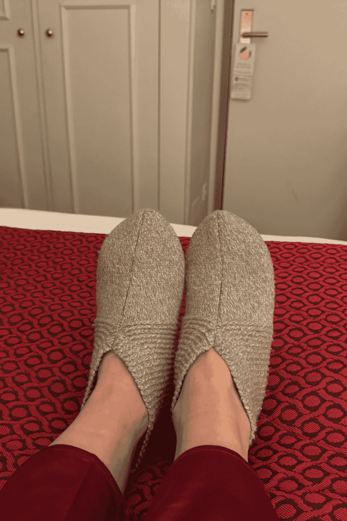 Kate relaxes on a red patterned hotel bed wearing cozy gray knit slippers and red pajama pants.