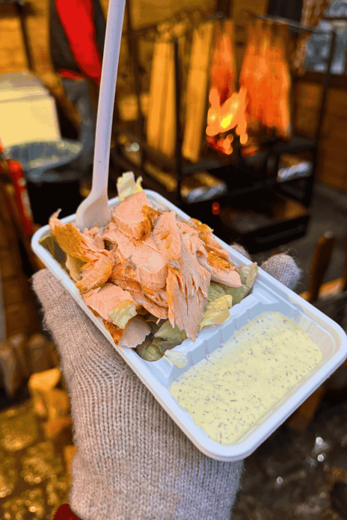 Gloved hand holding a tray of freshly flaked wood-fire grilled salmon on lettuce with dill sauce, served hot from a festive stall at the Brussels Christmas Market.

