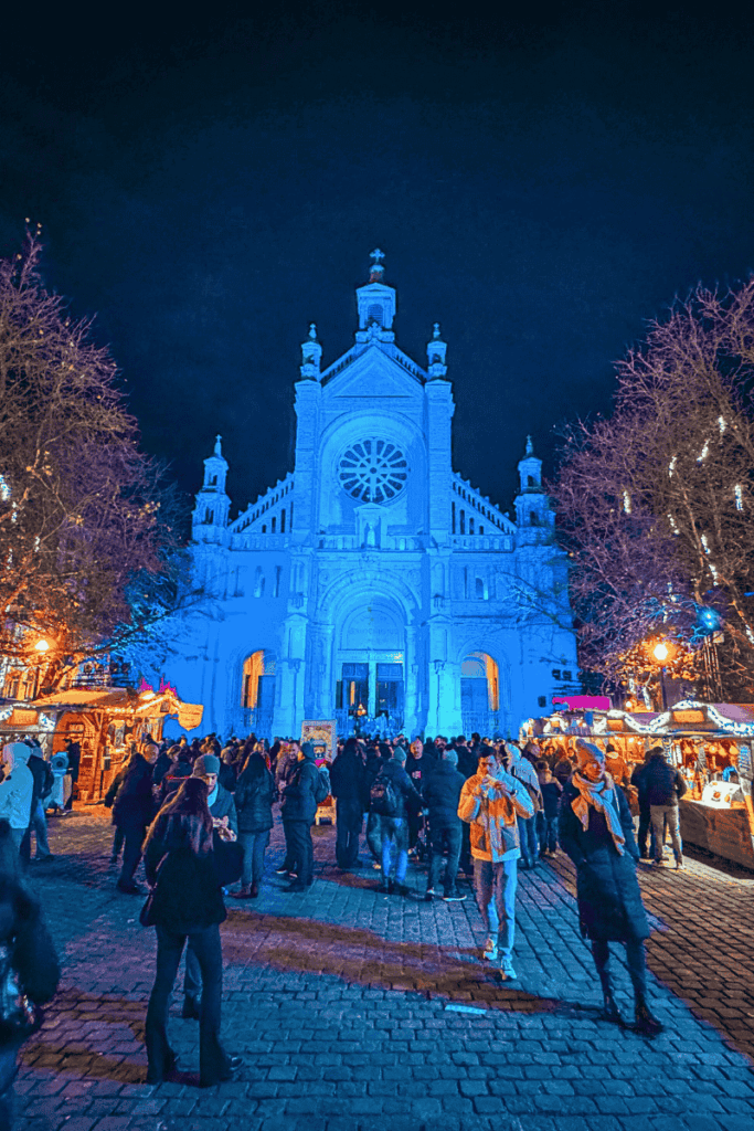 Crowds gather in front of the glowing blue-lit Sainte-Catherine Church, surrounded by festive Christmas market stalls and twinkling lights at night in Brussels.

