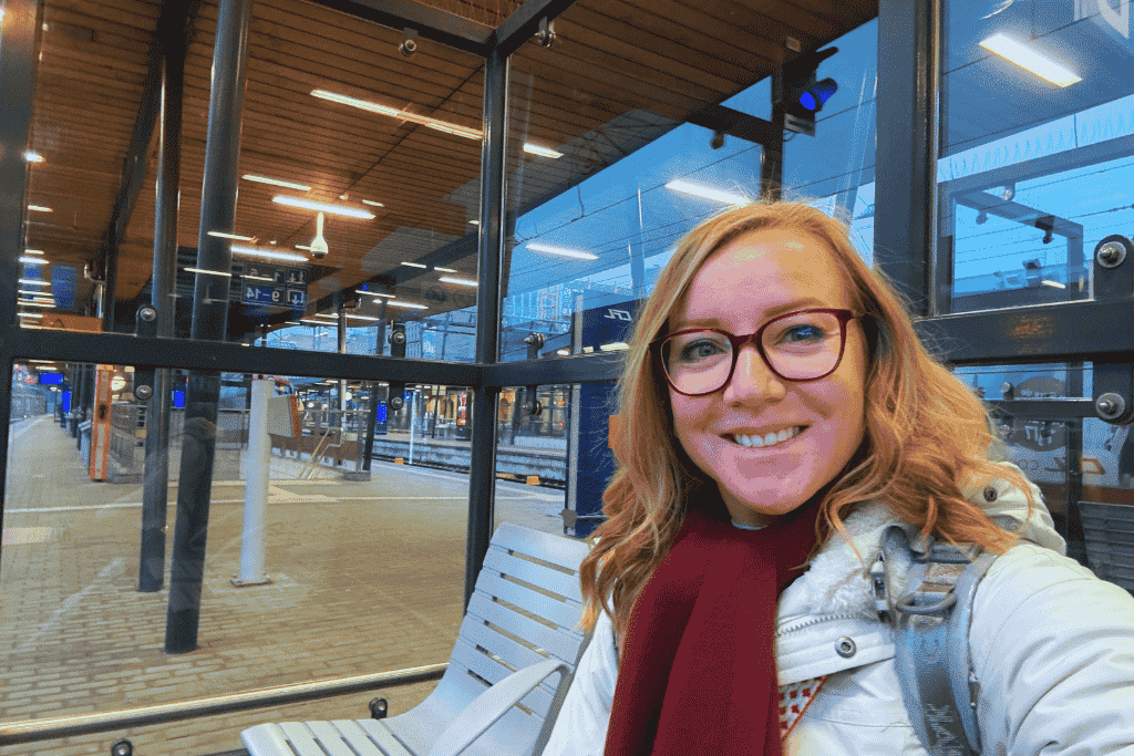 Kate smiles while sitting on a bench at a modern train station, surrounded by glass walls and overhead lighting.