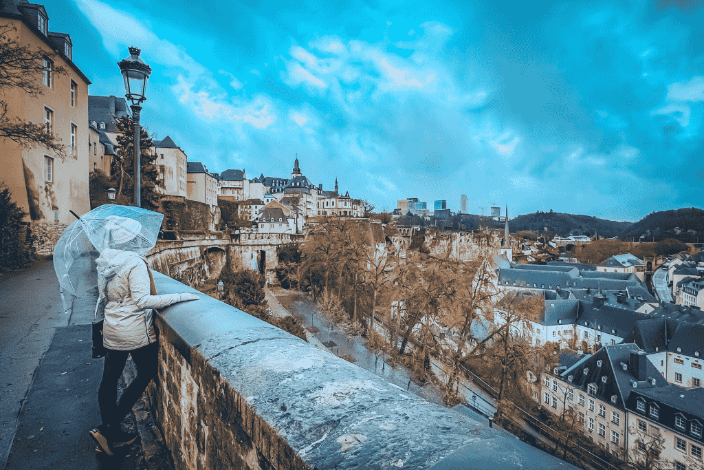 Kate leans on a stone wall with a clear umbrella, overlooking the dramatic valley and historic skyline of Luxembourg City under a moody sky.