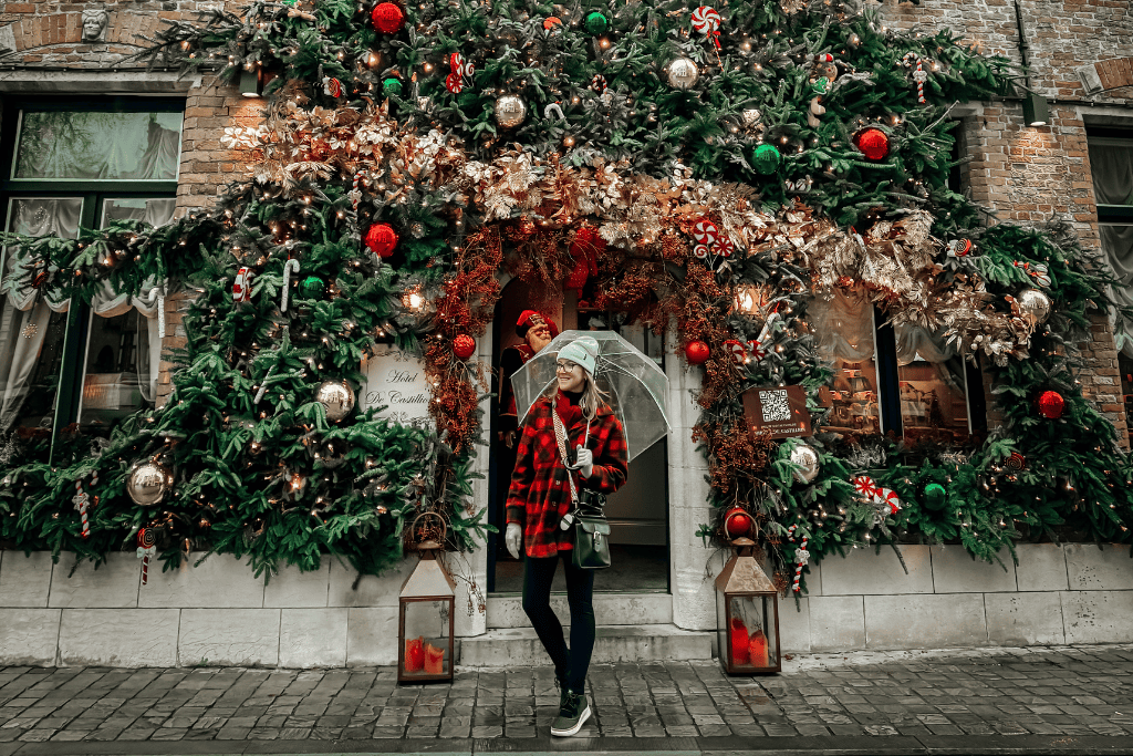 Kate stands under a clear umbrella in front of a hotel entrance overflowing with lush Christmas garlands, ornaments, and twinkling lights.