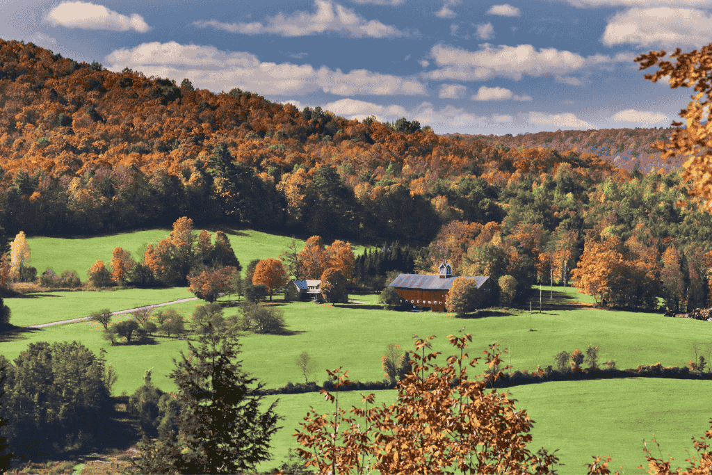 A peaceful Vermont farm sits in a wide green field surrounded by rolling hills covered in colorful fall foliage.