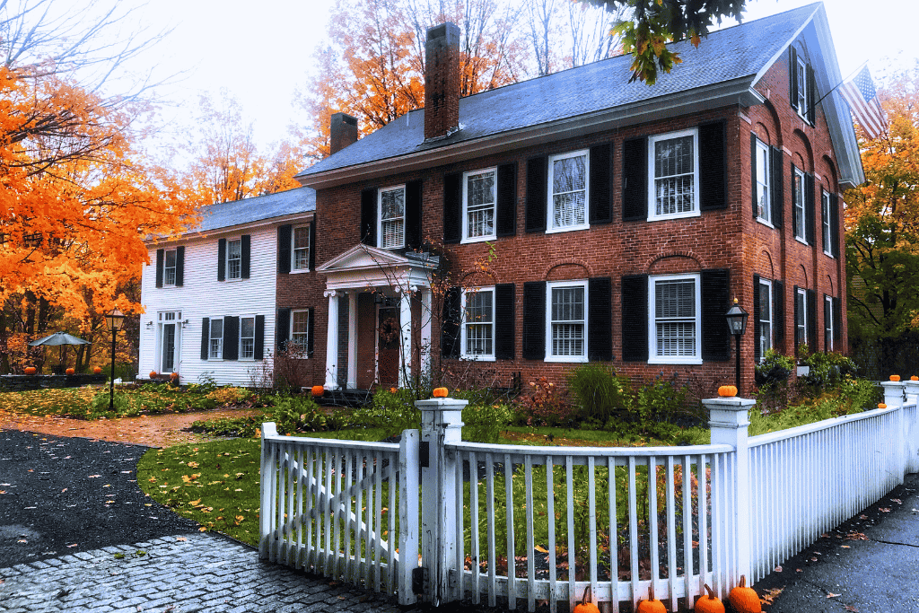 A colonial-style brick and white house in Woodstock, Vermont is trimmed with pumpkins and surrounded by bright fall trees.
