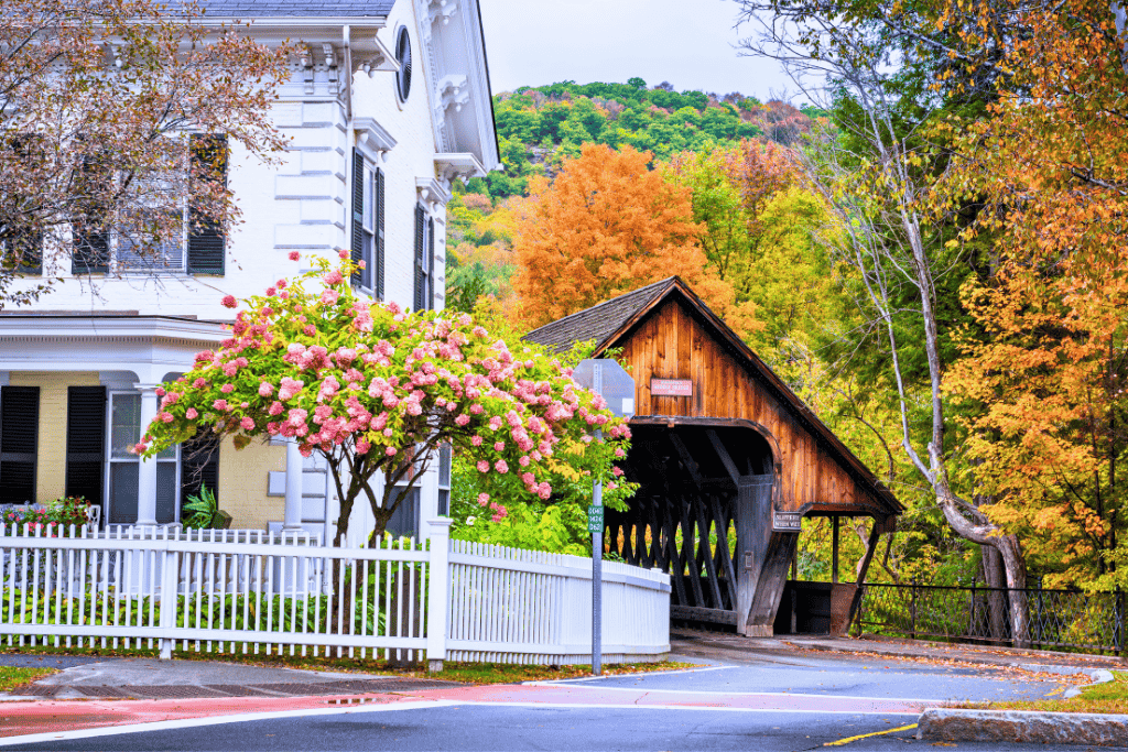 A charming covered bridge in Woodstock, Vermont stands next to a white Victorian house and a blooming pink tree, with colorful autumn hills behind.
