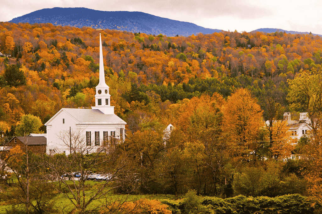 A white church with a tall steeple stands among vibrant autumn trees in a small Vermont town with forested hills rising behind.