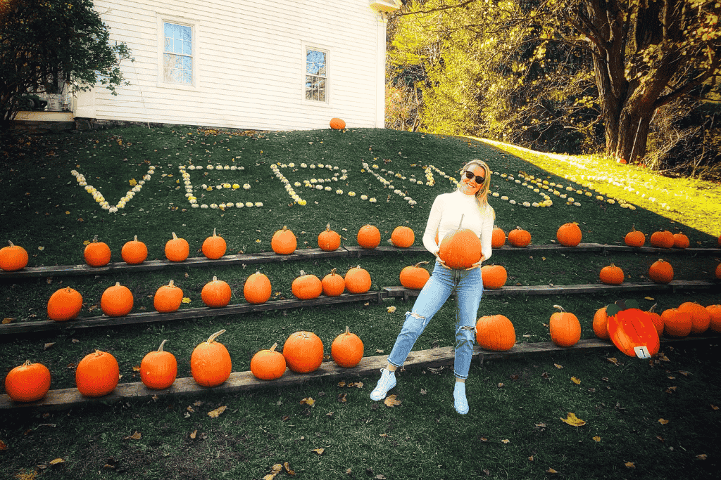 Kate stands smiling with a pumpkin in her hands at a festive display in Vermont, where rows of pumpkins line the grass and white gourds spell out “VERMONT” on a hillside.