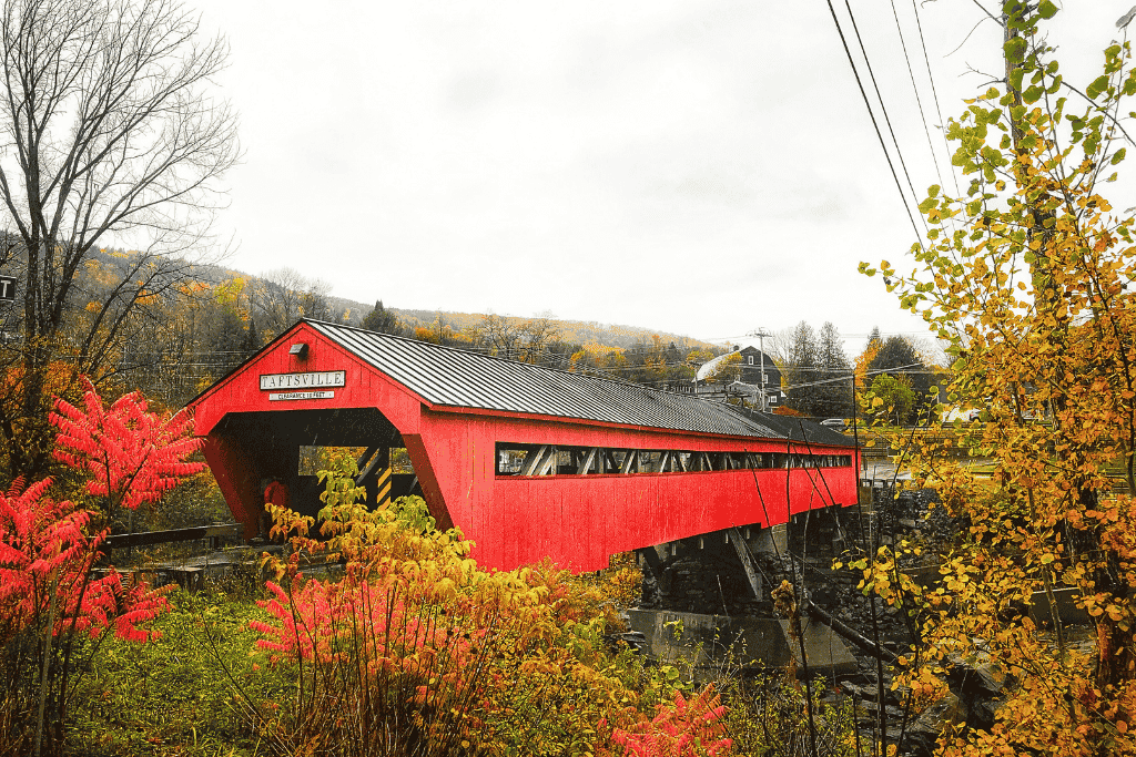 The bright red Taftsville Covered Bridge stretches over a rocky river, surrounded by vibrant fall foliage and a small Vermont village in the distance.