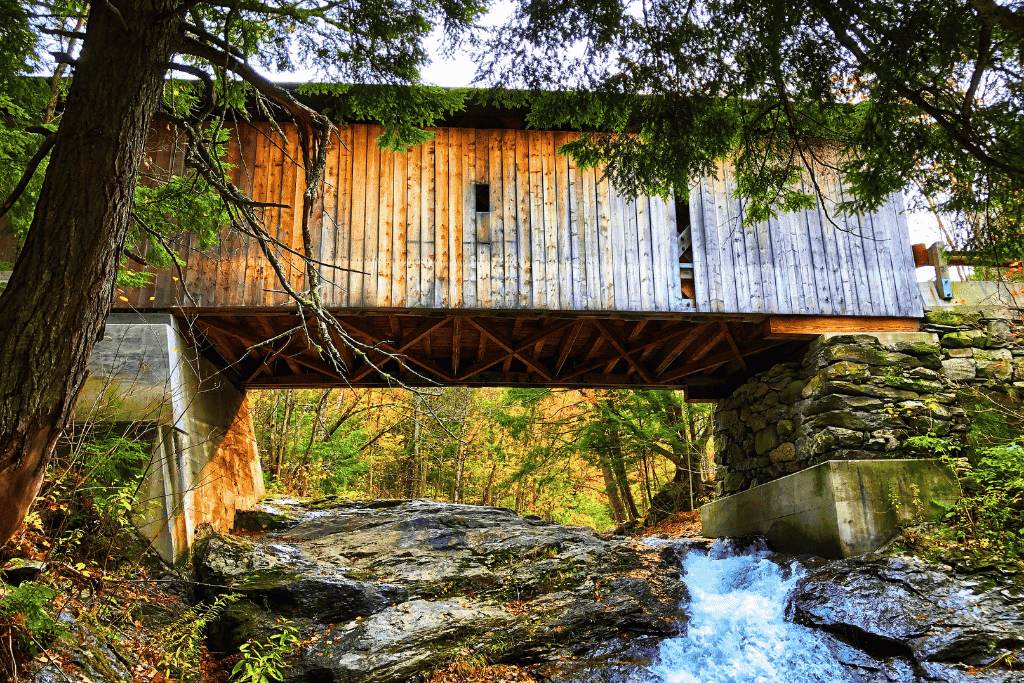 A wooden covered bridge crosses over a rocky stream with water rushing below in a forested Vermont setting.