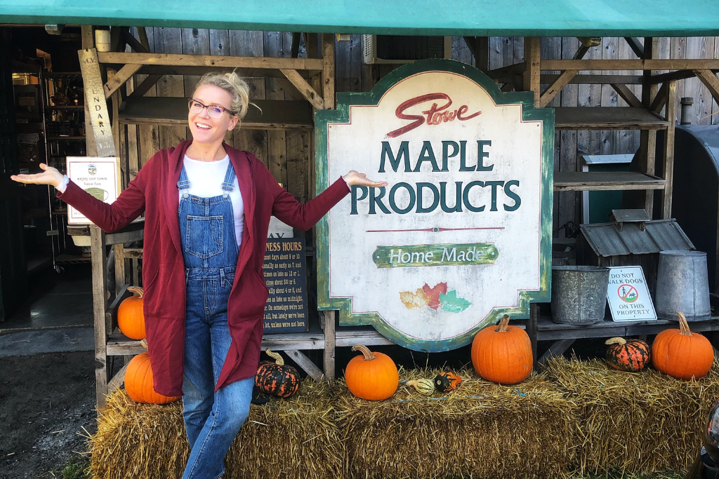 Kate stands smiling with arms outstretched in front of a rustic "Stowe Maple Products" sign surrounded by pumpkins and hay bales in Vermont.