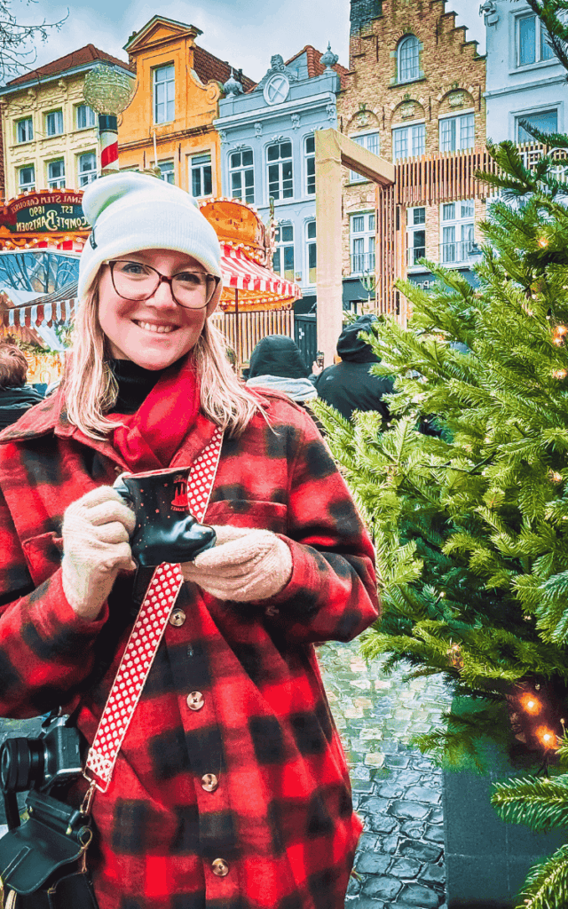 Kate holds a boot-shaped mug of festive drink beside a decorated Christmas tree at the Bruges Christmas Market.