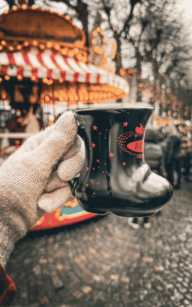A gloved hand holds a black boot-shaped Bruges Christmas Market mug in front of a glowing carousel.
