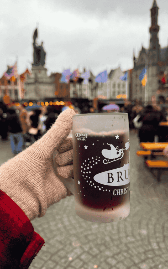 A gloved hand holds a frosted Bruges Christmas Market mug filled with mulled wine against the backdrop of Markt Square.