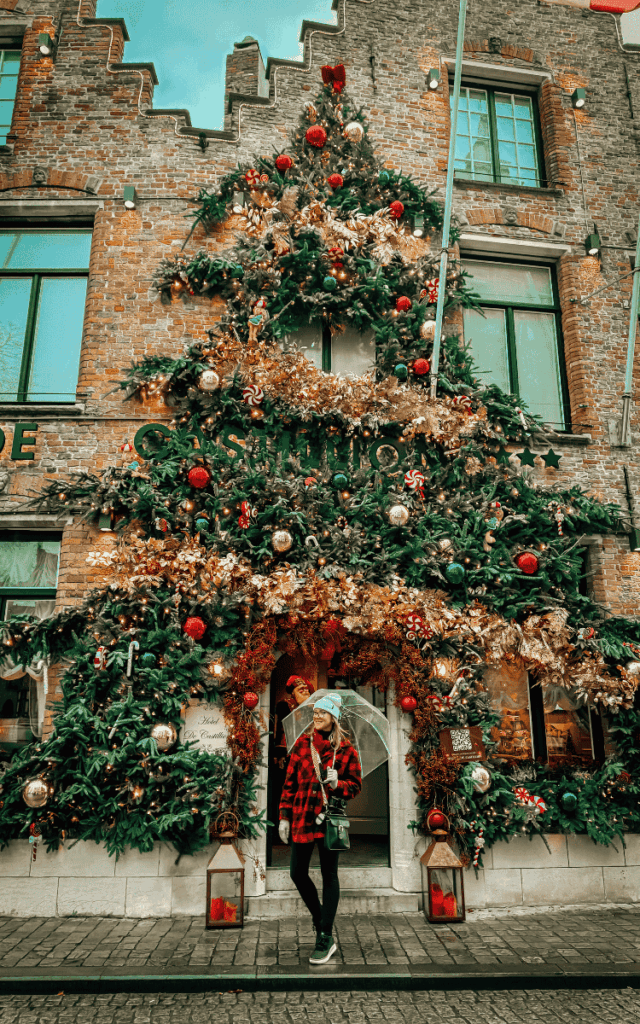 Kate holds a clear umbrella beneath an elaborate Christmas tree display covering the façade of a Bruges building.
