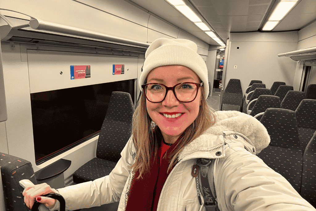 Kate smiles for a selfie inside an empty train carriage, wearing a white beanie, glasses, and a light winter coat