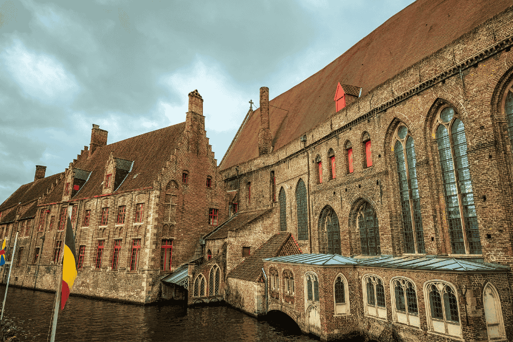 The medieval brick buildings of Bruges line a canal under a cloudy winter sky, with Belgian flags in the foreground.