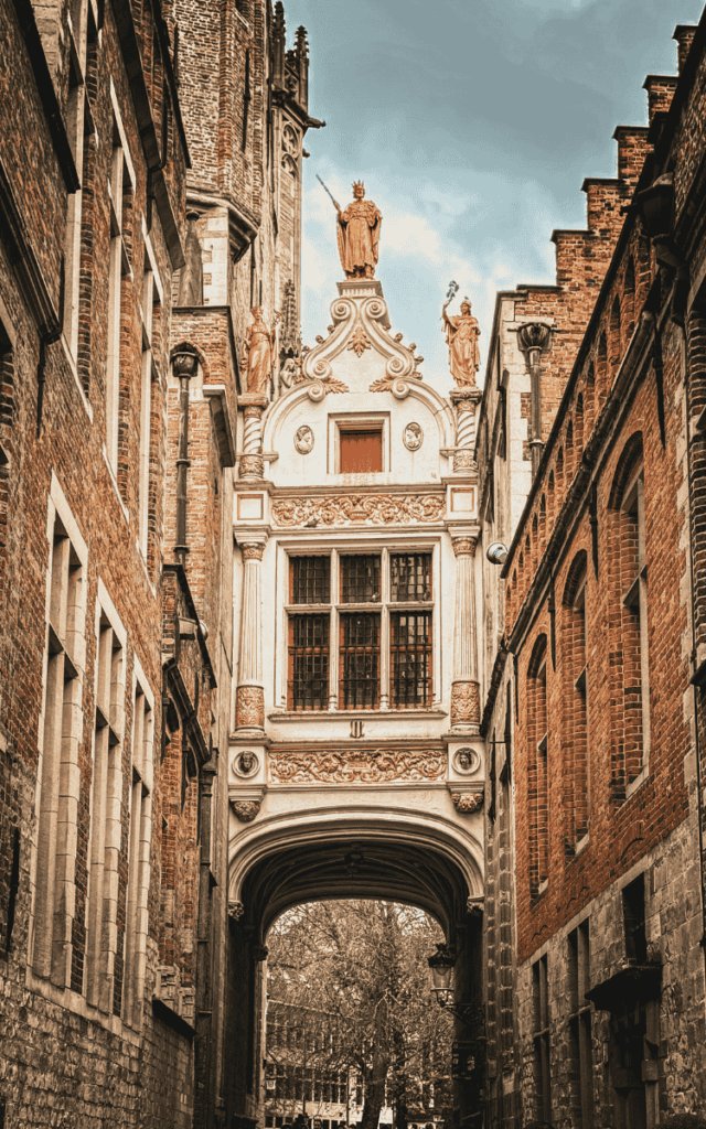 An ornate arched passage in Bruges is topped with statues and decorative carvings between historic brick buildings.
