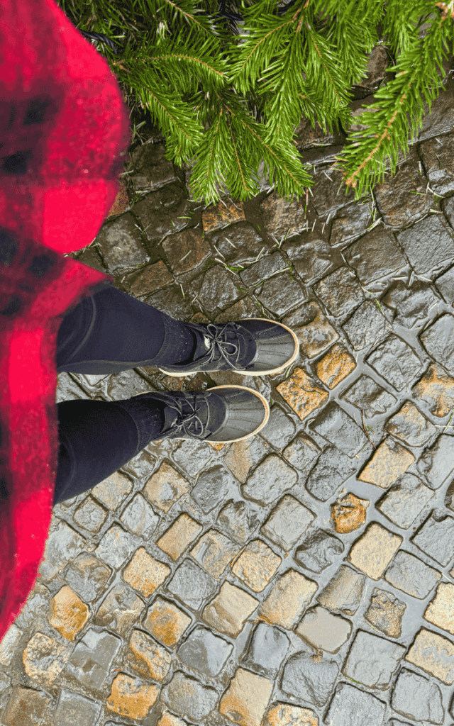 Kate stands on wet cobblestones beside pine branches, wearing black boots and a red plaid coat.
