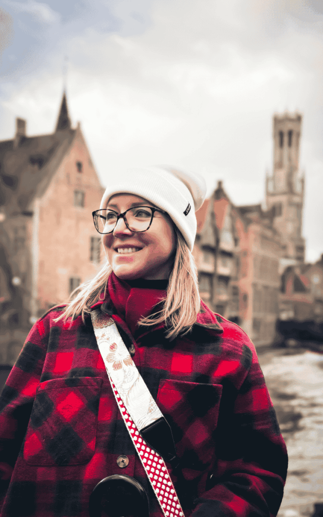 Kate smiles in a white beanie and red plaid coat with Bruges’ historic canal buildings blurred in the background.