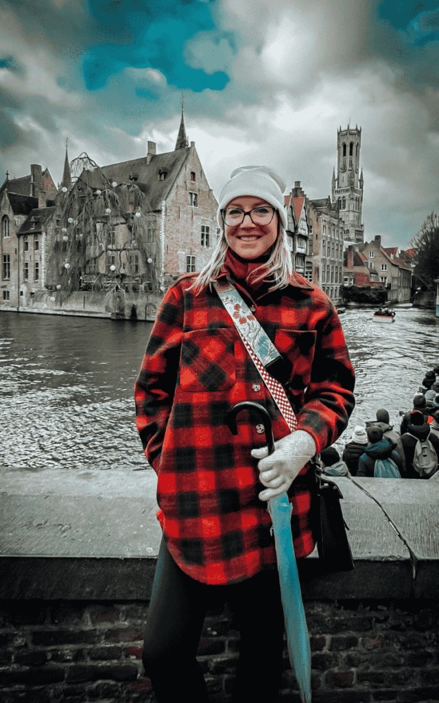 Kate stands by a canal in Bruges holding a blue umbrella, with the Belfry tower and medieval buildings behind her.