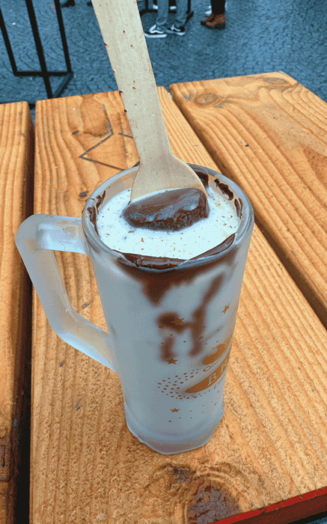 A frosted Bruges Christmas Market mug holds creamy hot chocolate topped with a wooden spoon coated in melted chocolate.