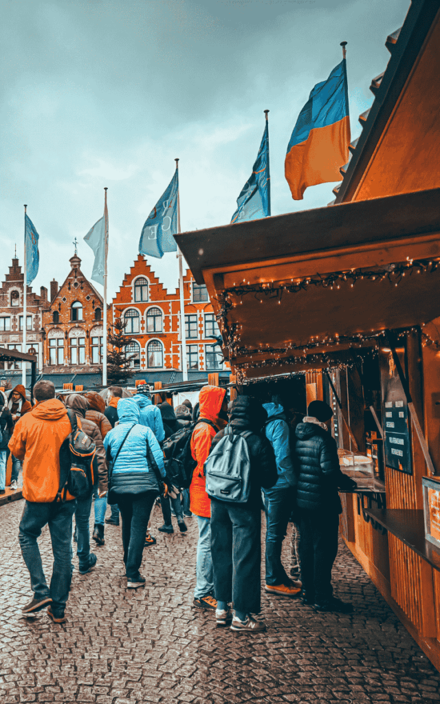 People in colorful winter coats line up at a wooden stall in the Bruges Christmas Market, with flags and gabled buildings in the background.