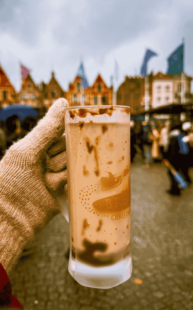 A gloved hand holds a Bruges Christmas Market mug filled with creamy hot chocolate against a blurred festive square backdrop.