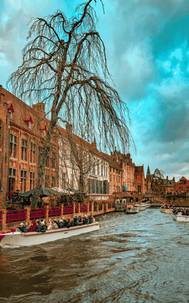 A tour boat filled with passengers glides along Bruges’ canal past historic brick buildings and a leafless tree.