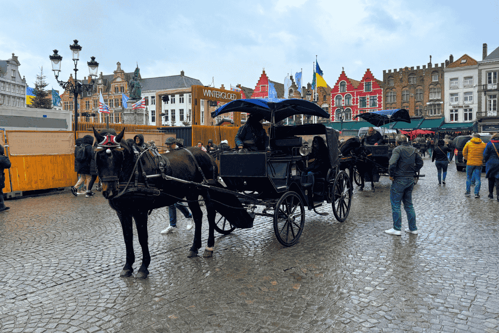 Horse-drawn carriages wait in Bruges’ Markt Square with colorful gabled buildings and winter flags in the background.