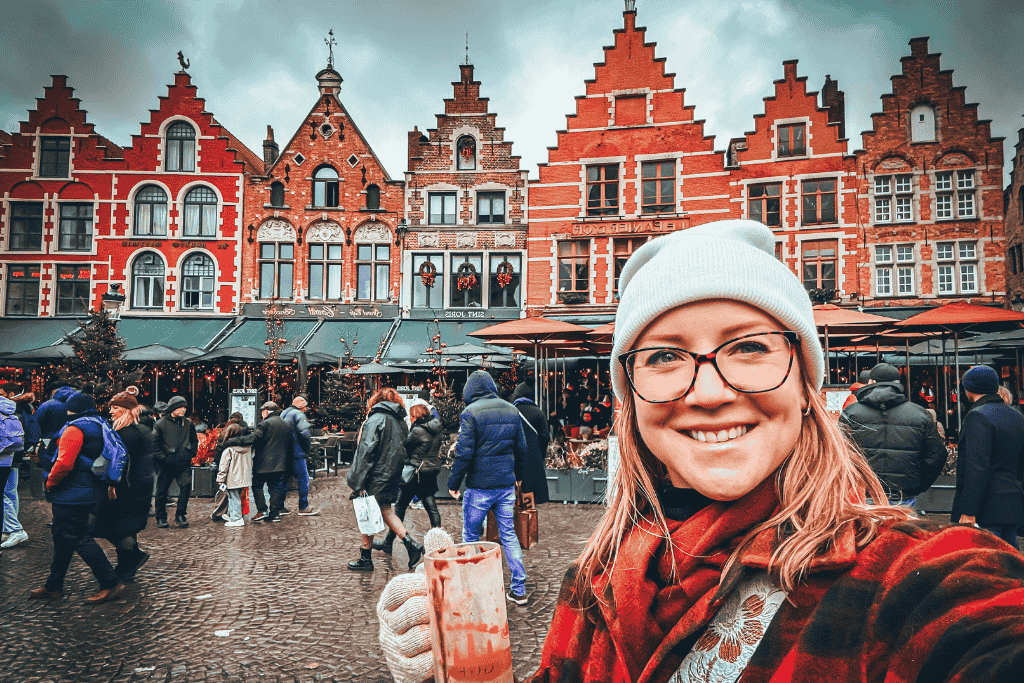 Kate smiles while holding a festive drink in front of the red-brick gabled buildings at the Bruges Christmas Market.