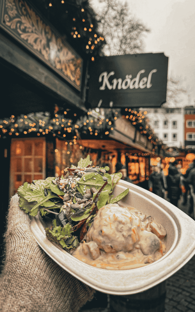 A gloved hand holds a plate of creamy mushroom sauce knödel with mixed greens in front of a festive “Knödel” food stall at the Cologne Christmas Market.