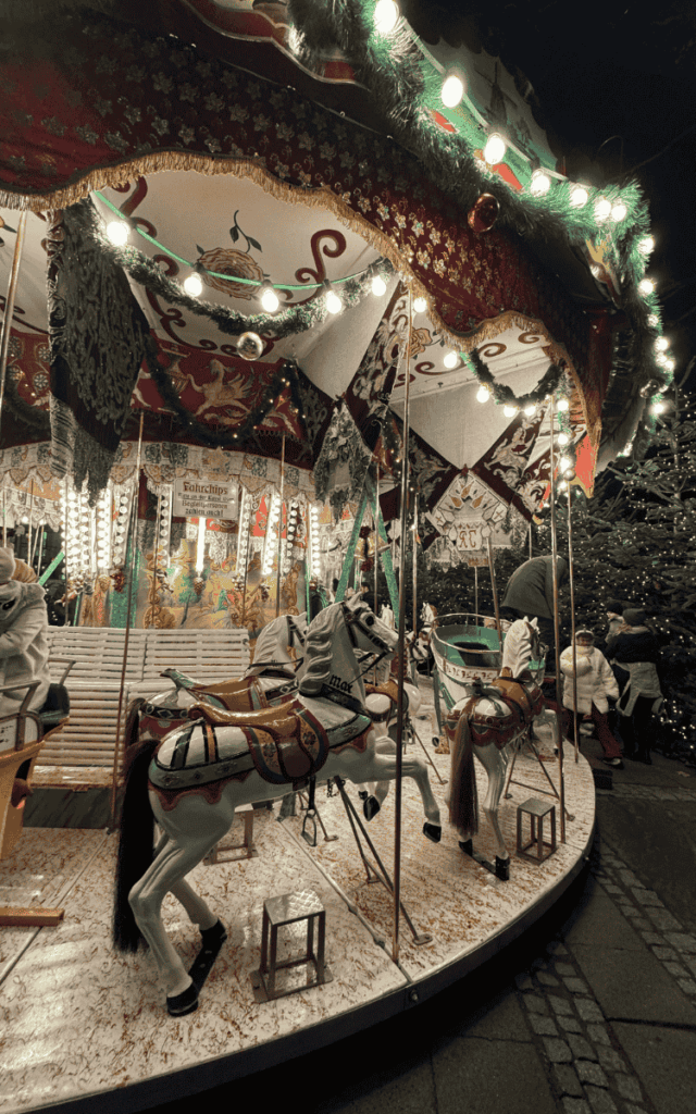 A vintage carousel decorated with festive garlands and lights features painted white horses and wooden benches at the Cologne Christmas Market.