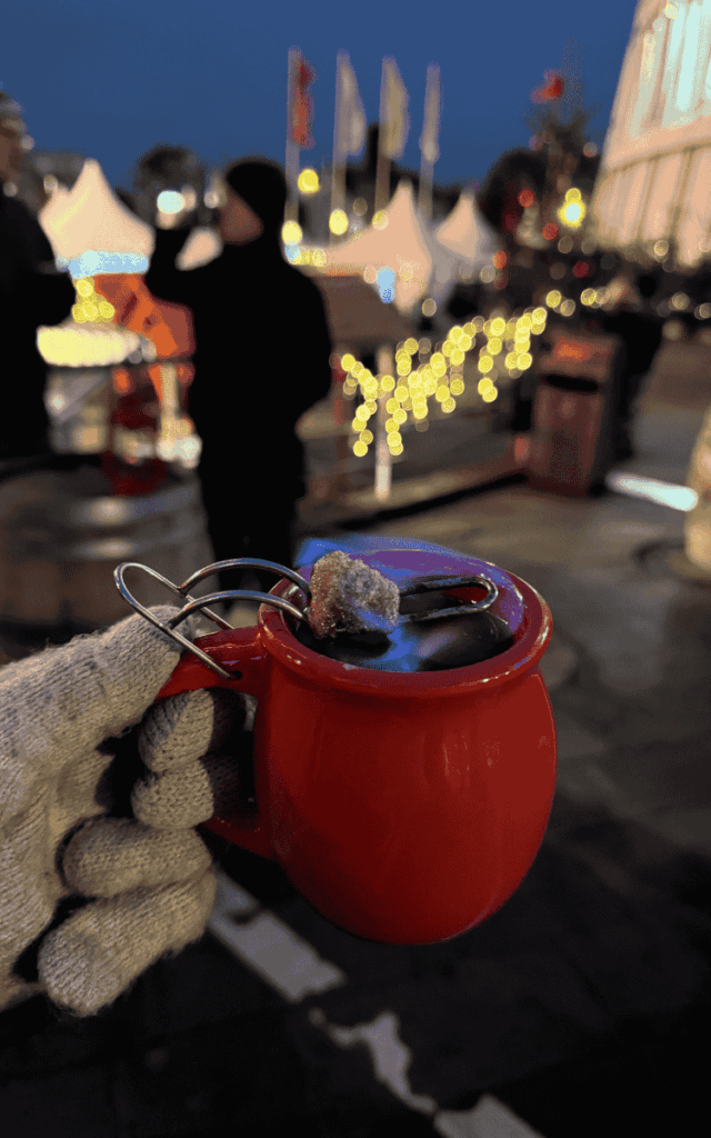 A gloved hand holds a red mug of flaming Feuerzangenbowle topped with a burning sugar cube at the Cologne Christmas Market.